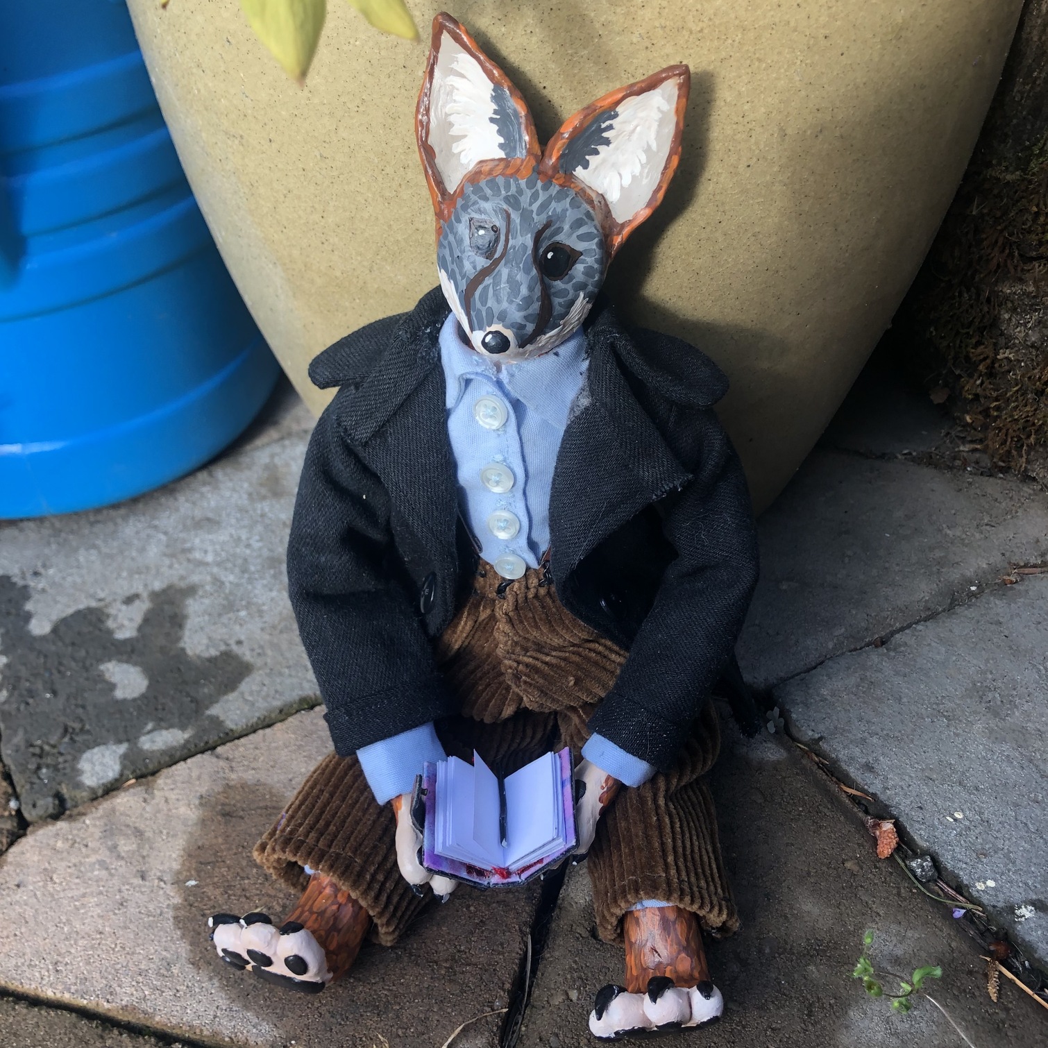 A small humanoid fox doll with clay face and paws, dressed in a wool coat, sits in a garden with a book. Link takes you to project page.