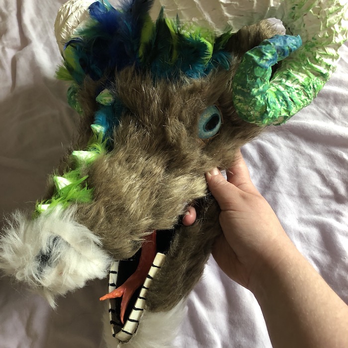 A cardboard mask of an intimidating gost monster with large white horns, the fur is brown and there are blue and green feathers circling each horn. Link takes you to project page.