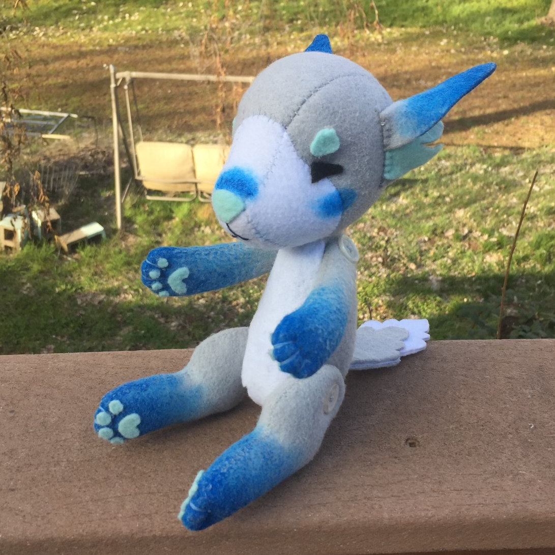 A grey fox plush with a white belly, the eyes are squinted shut and the ends of the paws are painted blue. Picture links to project page.