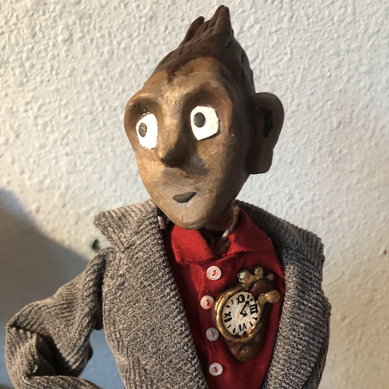 A small humanoid puppet from the waist-up. His skin is the texture of wood and he wears a red collared shirt and grey coat. On his chest is a clock shaped like a heart. Picture links to project page.