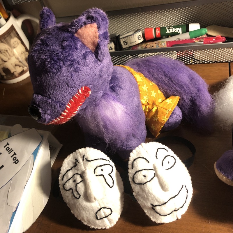A sitting purple dog plush with no face, wearing orange shorts. In front of him are two white felt masks showing scared and an excited expressions. Picture links to project page.