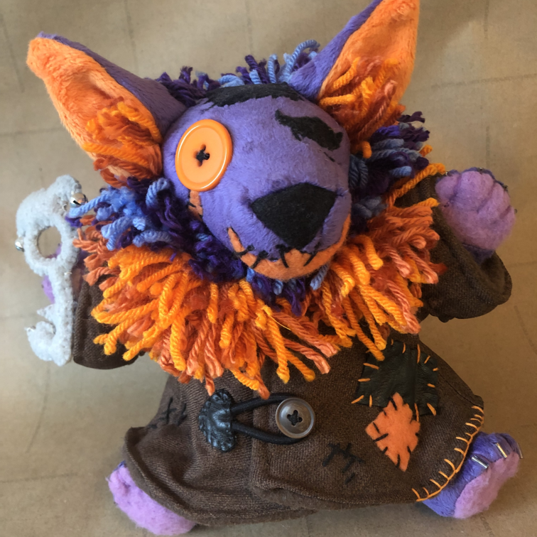 A plush of a purple cat creature with fluffy yarn fur, they wear a patchy brown coat with a fluffy orange ruff. Picture links to project page.